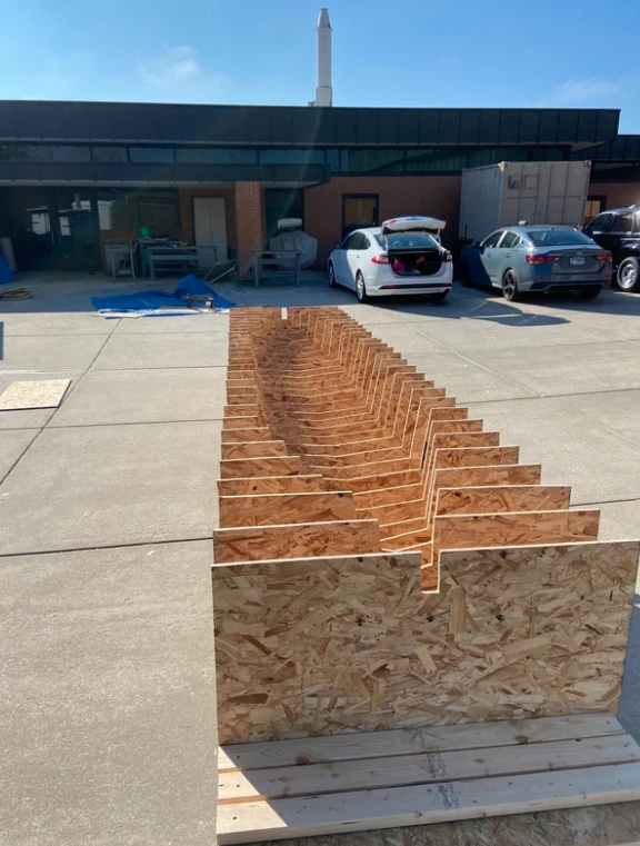 Wooden panels arranged vertically in rows on a concrete surface outside, near parked cars and buildings—an installation reminiscent of methods endorsed by the American Society of Civil Engineers.